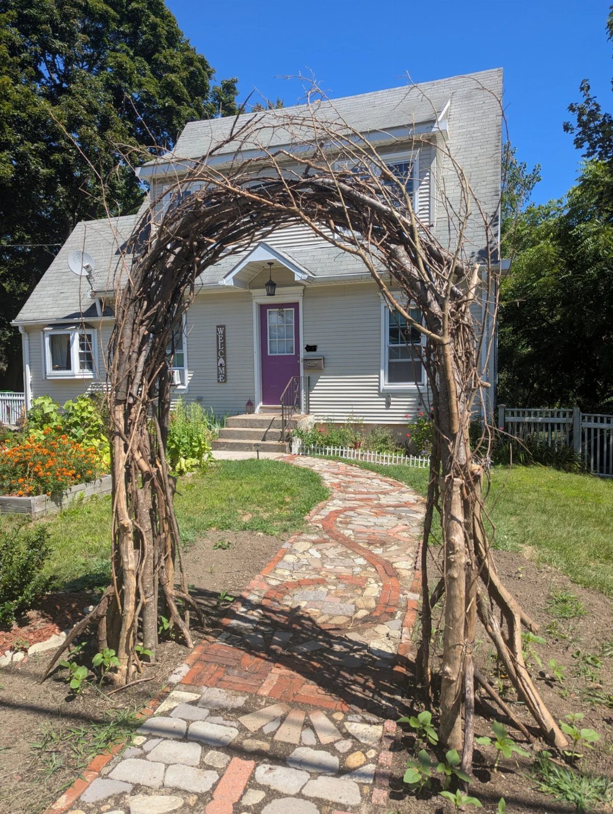 A rustic branch archway frames a stone pathway leading toward a white house.