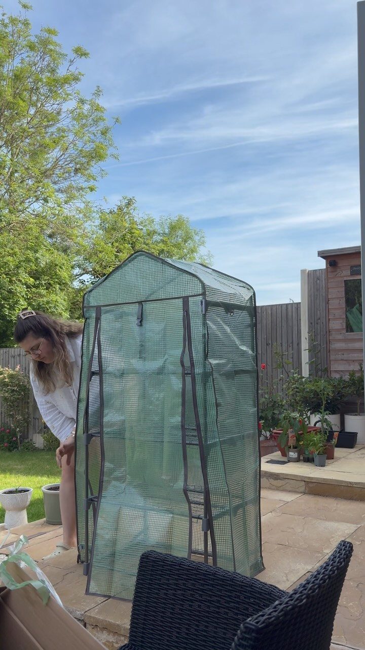 A woman stands beside a small green portable greenhouse kit on a stone patio.
