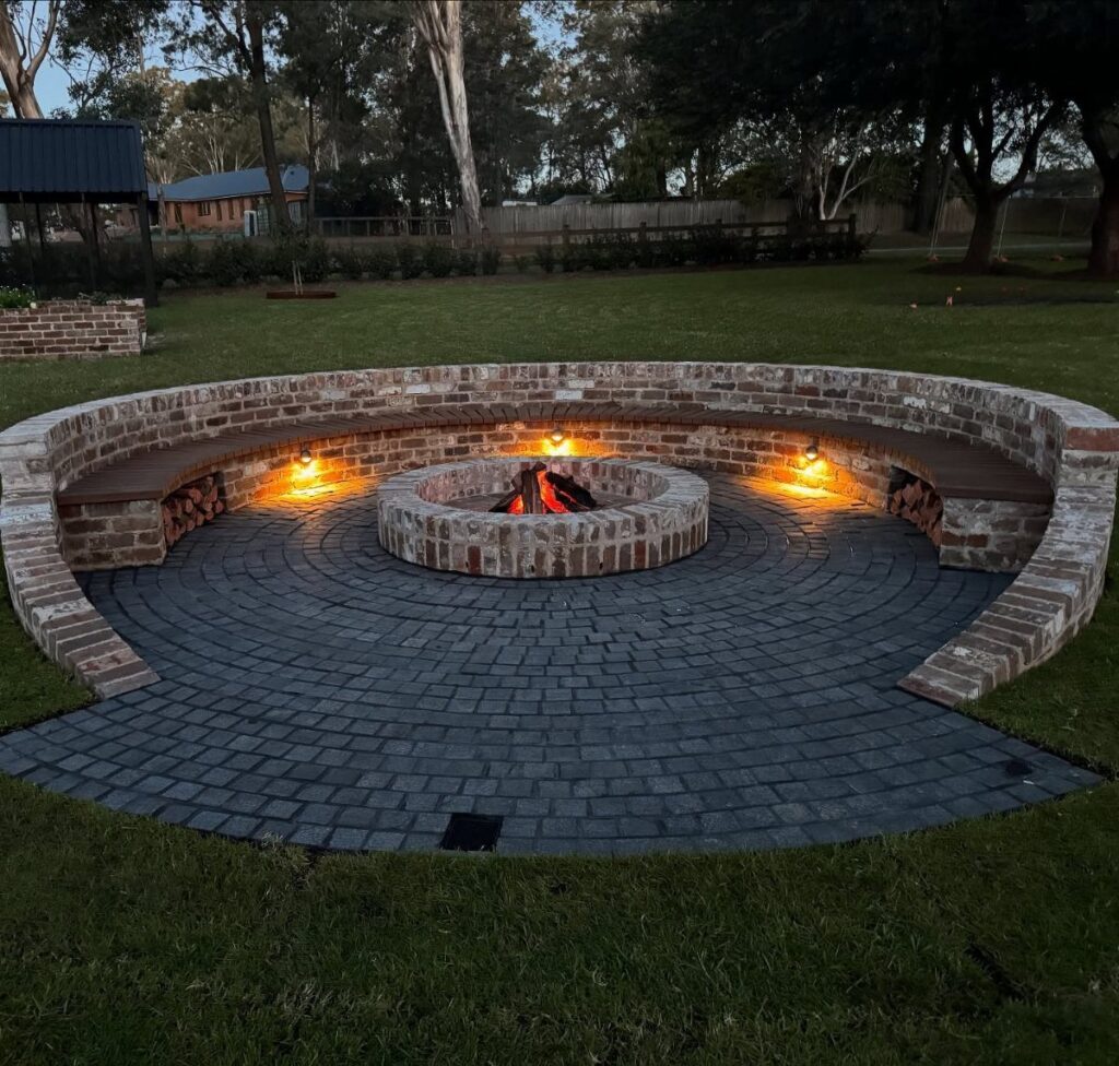 A circular brick fire pit with a glowing fire surrounded by a curved brick bench and patio.