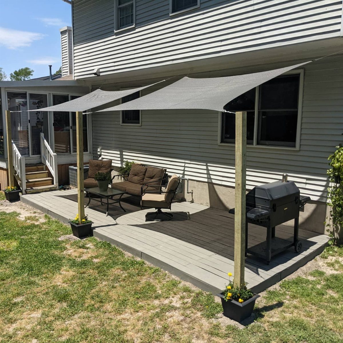 Grey shade sails stretched over a wooden deck with outdoor furniture and a grill.
