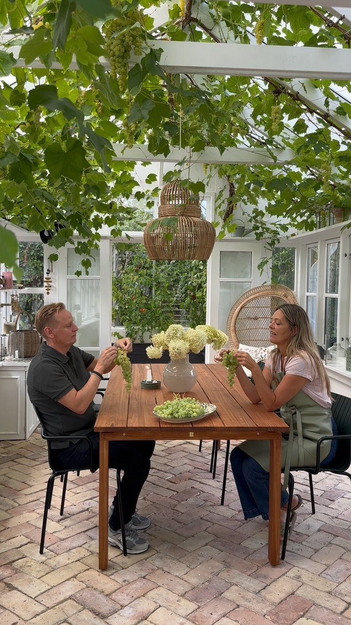 A man and woman sit at a wooden table arranging flowers under a vine-covered pergola.