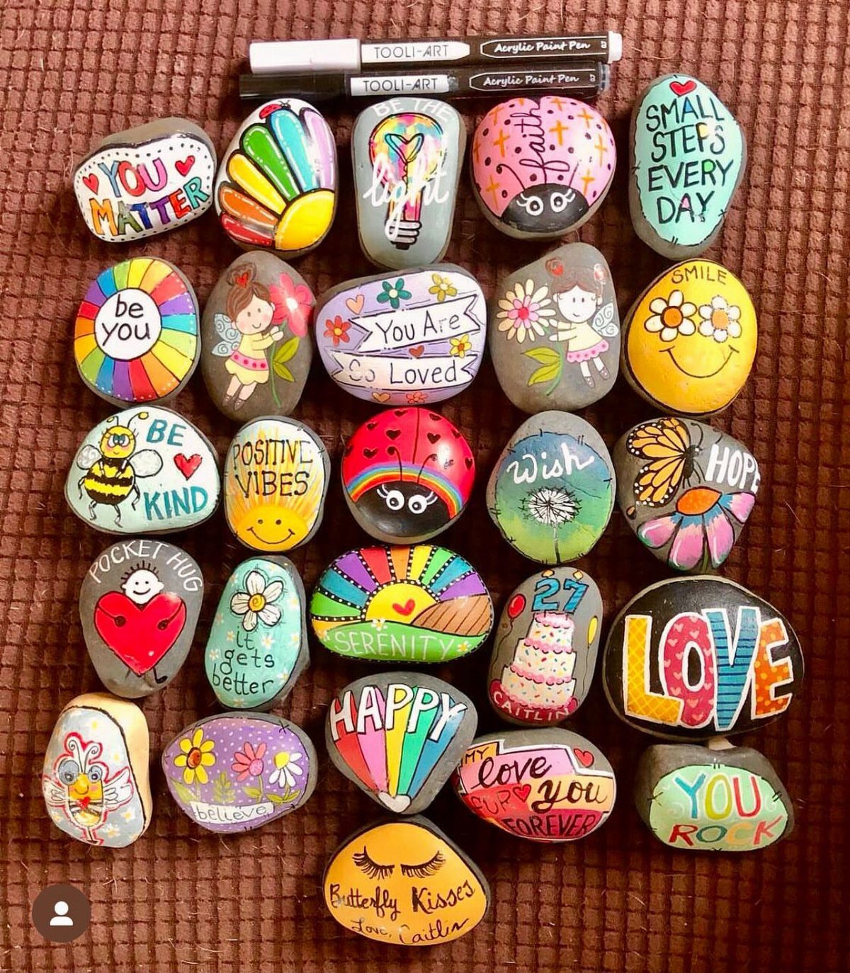 Many colorful hand-painted rocks with inspirational words and designs arranged on a textured brown surface.