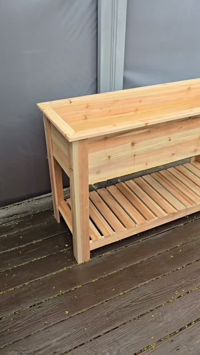 A wooden elevated garden planter box featuring a slatted storage shelf underneath on a deck.