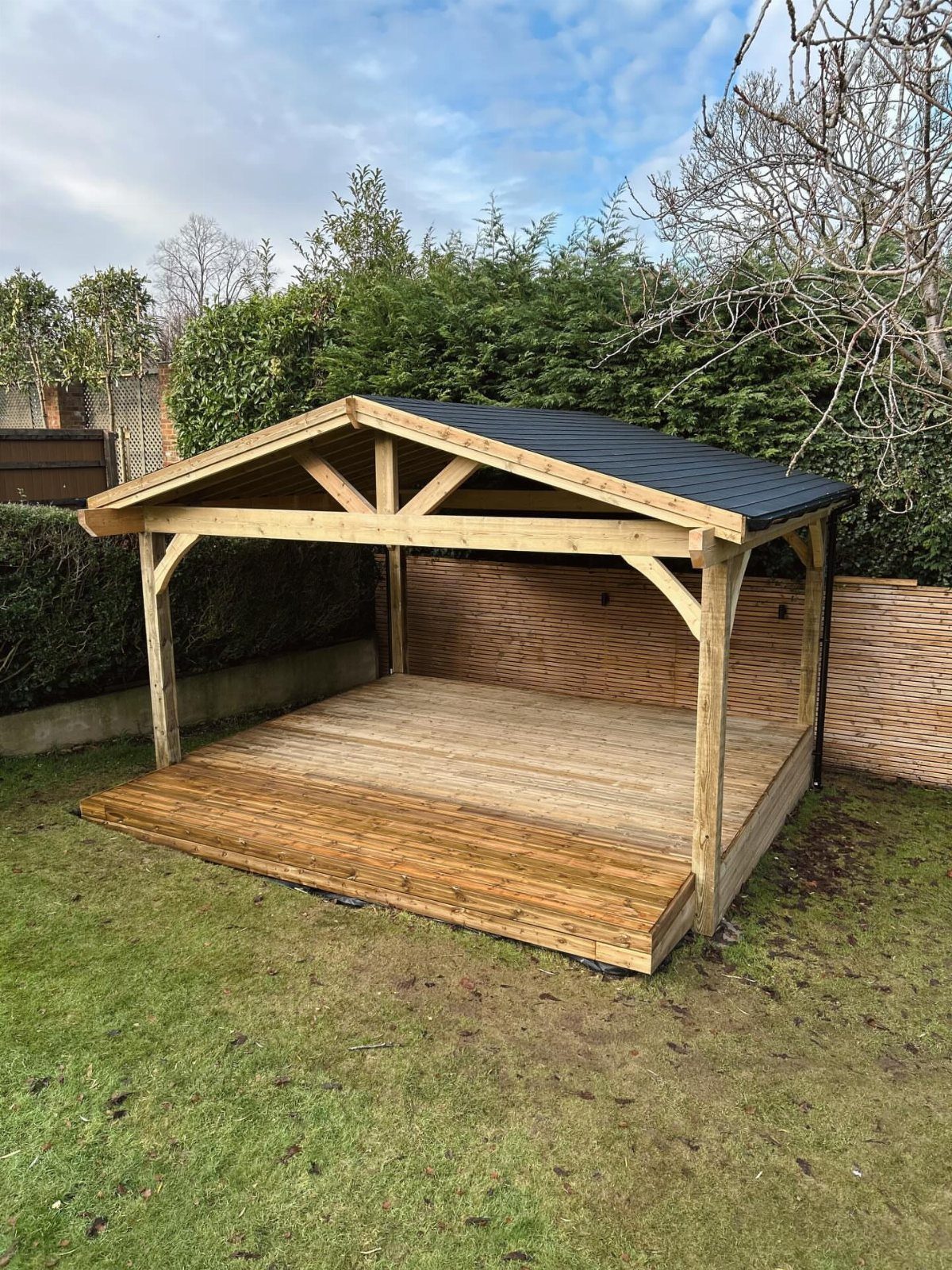 A wooden gazebo with a dark shingled roof built on a deck in a garden.
