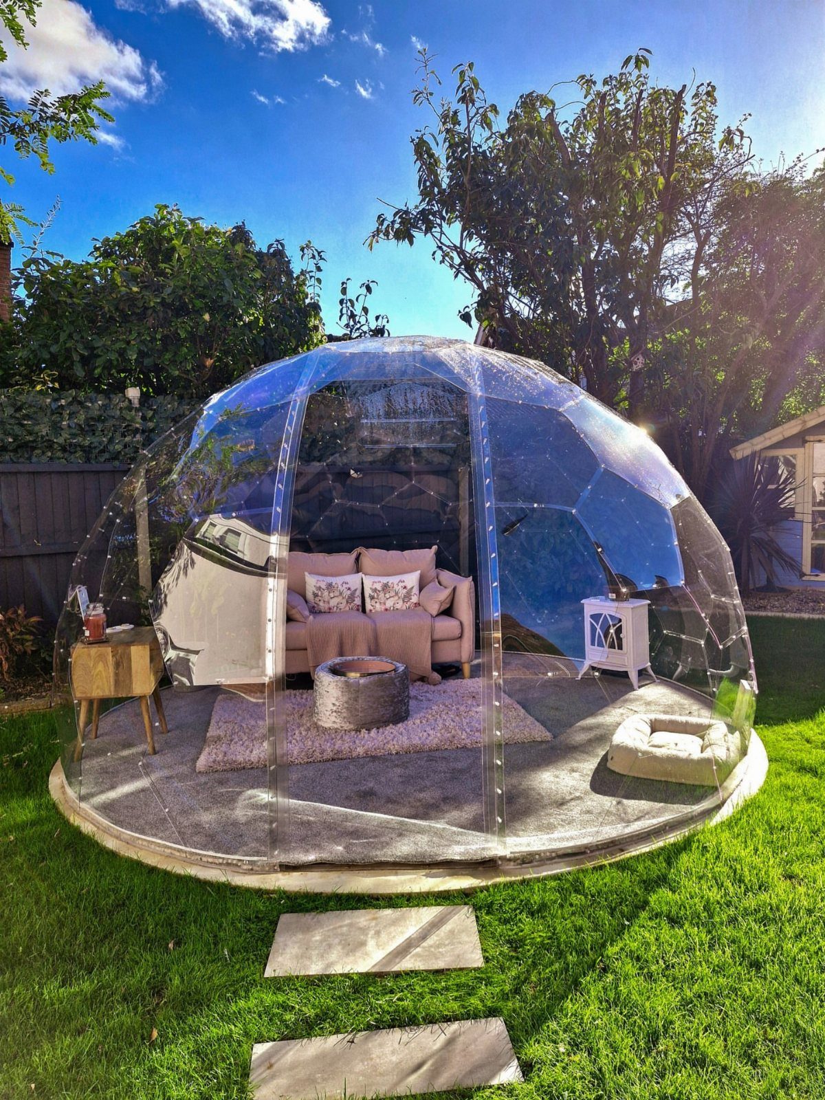 A transparent geodesic dome garden structure containing a cozy outdoor living room setup on a lawn.