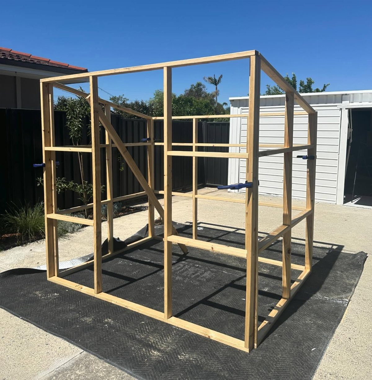 A wooden greenhouse frame structure sits on a black foundation mat outdoors.