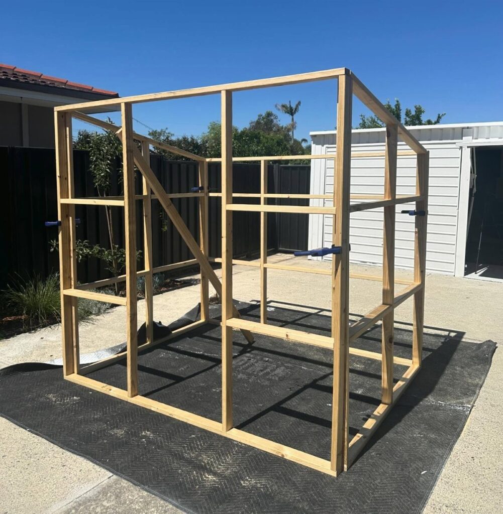 A wooden greenhouse frame structure sits on a black foundation mat outdoors.