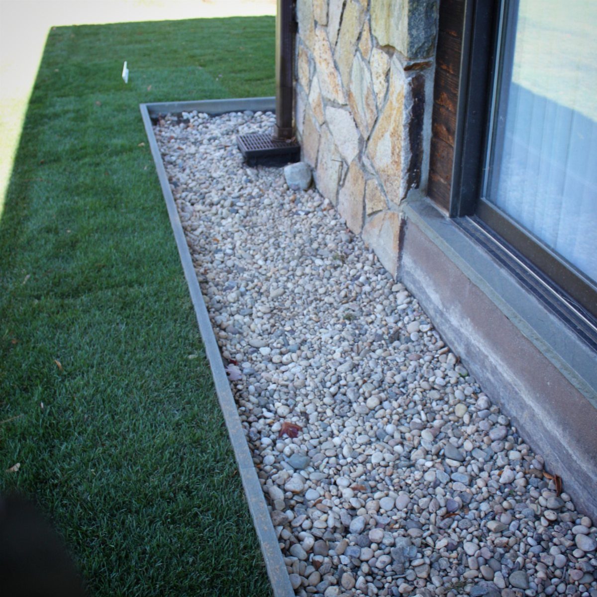 A narrow gravel border with metal edging runs along a stone house foundation next to green grass.