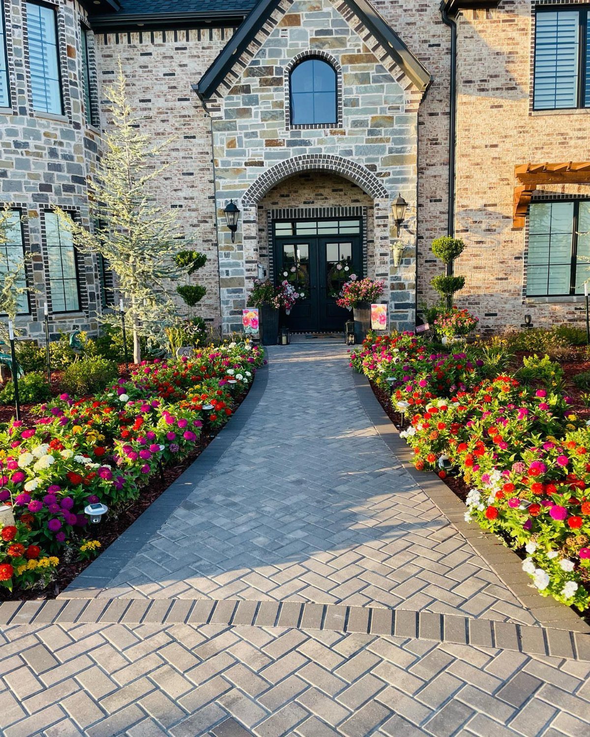 Colorful flower beds line a brick walkway leading to a stone house entrance.