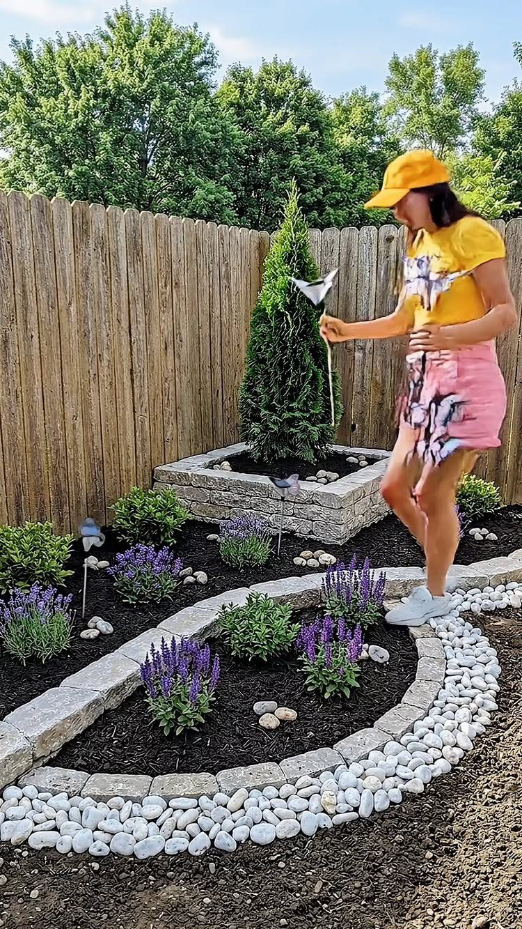 Backyard with Trees: 10 Layouts for Shade, Privacy and Outdoor Living 5 A woman tending to a landscaped garden bed with dark mulch and purple flowers.