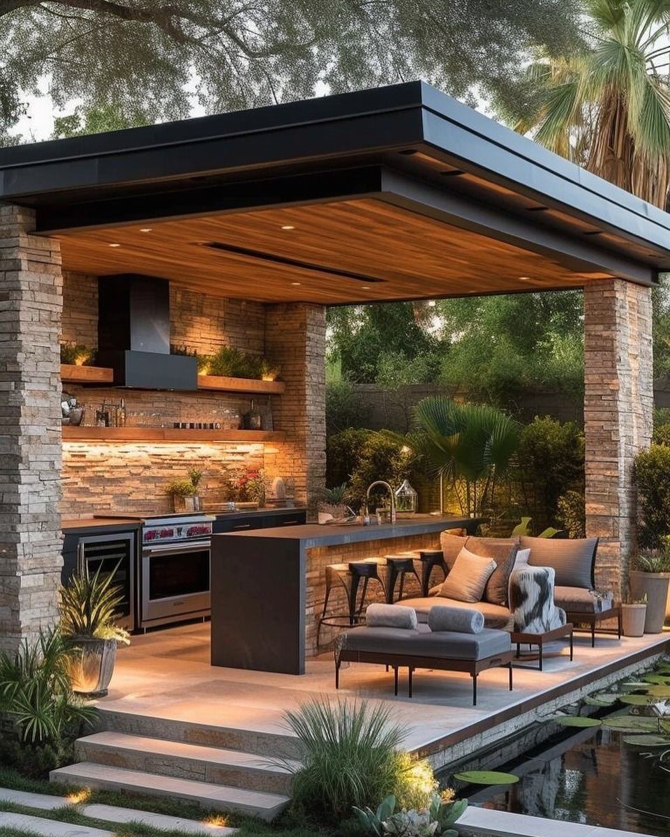 A modern outdoor kitchen with stone pillars, wooden ceiling, and lounge seating by a pond.