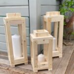 Three handmade wooden lanterns with white pillar candles sitting on a wooden deck outdoors.