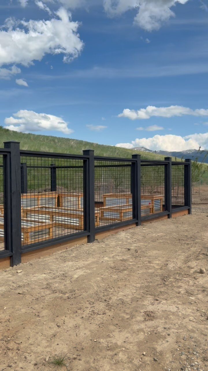 Wooden raised garden beds enclosed by a black metal wire mesh fence in a rural landscape.