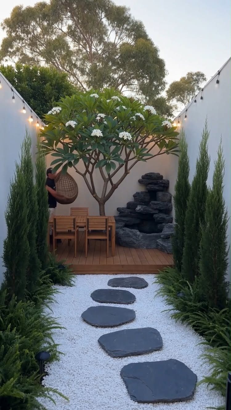 A small wooden deck with dining furniture, a stone water feature, and a white gravel path.