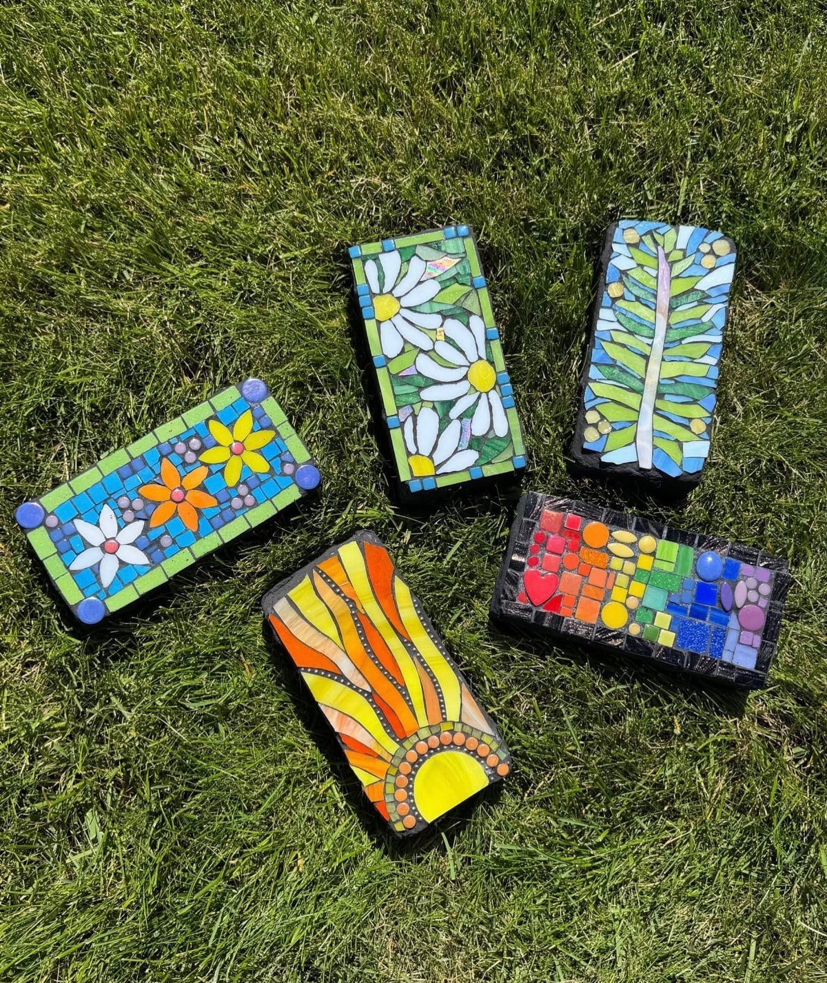 Five colorful mosaic stepping stones with floral and sun patterns resting on green grass.