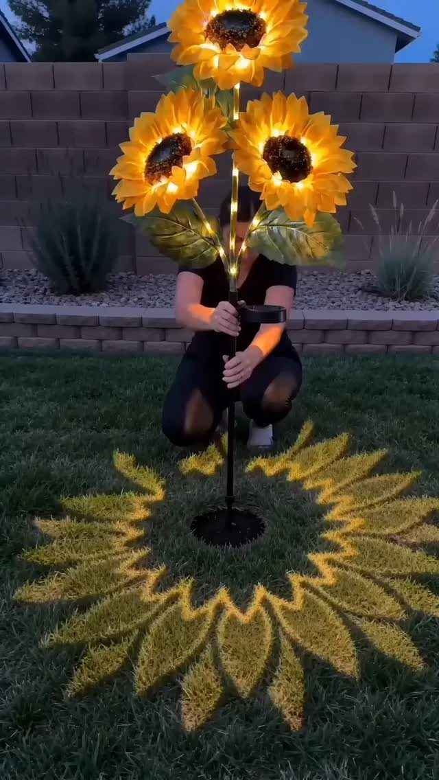 Three glowing sunflower solar lights on a black pole in a grassy yard.