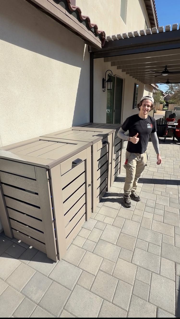 12 Side House Landscaping Ideas for Narrow Spaces 6 A man standing beside two tan wooden slat trash bin enclosures on a stone patio.