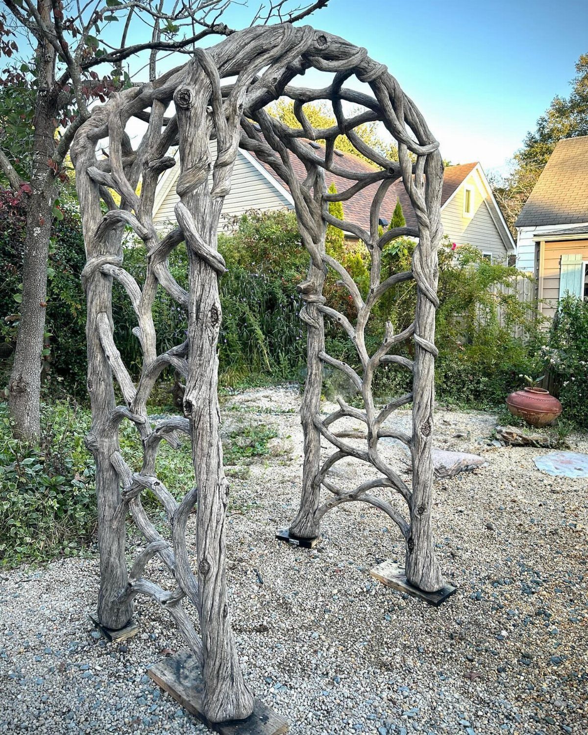 An arched garden trellis made from intertwined, weathered grey tree branches standing on gravel.