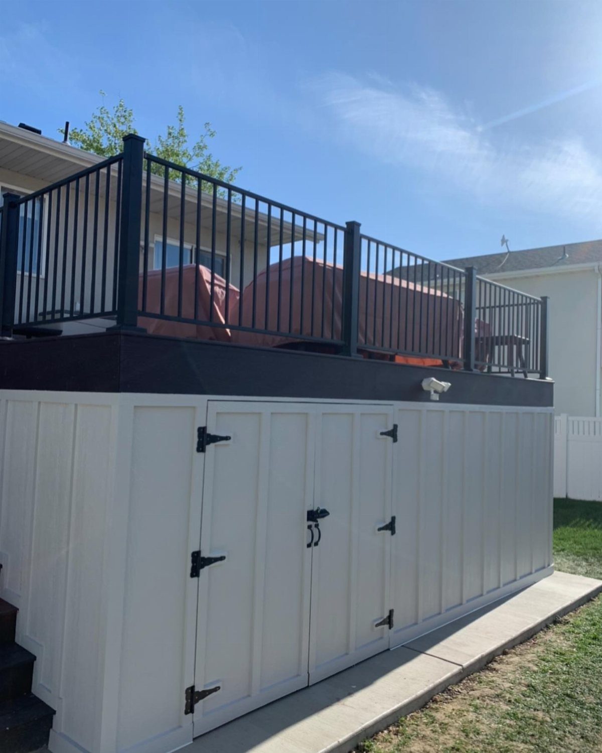 A white storage shed constructed underneath an elevated wooden deck with black metal railings.