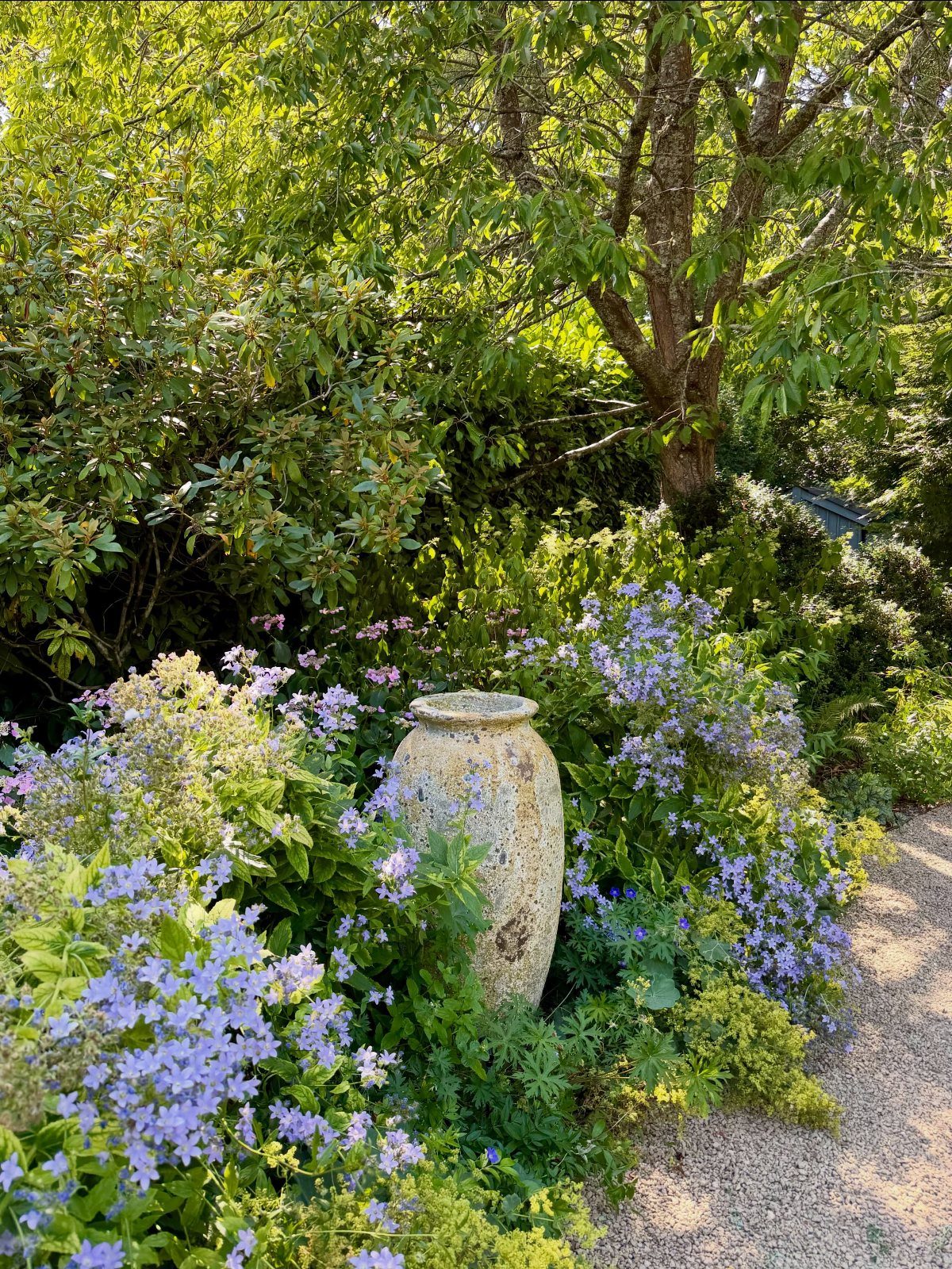 A large stone urn surrounded by purple wildflowers and green bushes next to a gravel path.